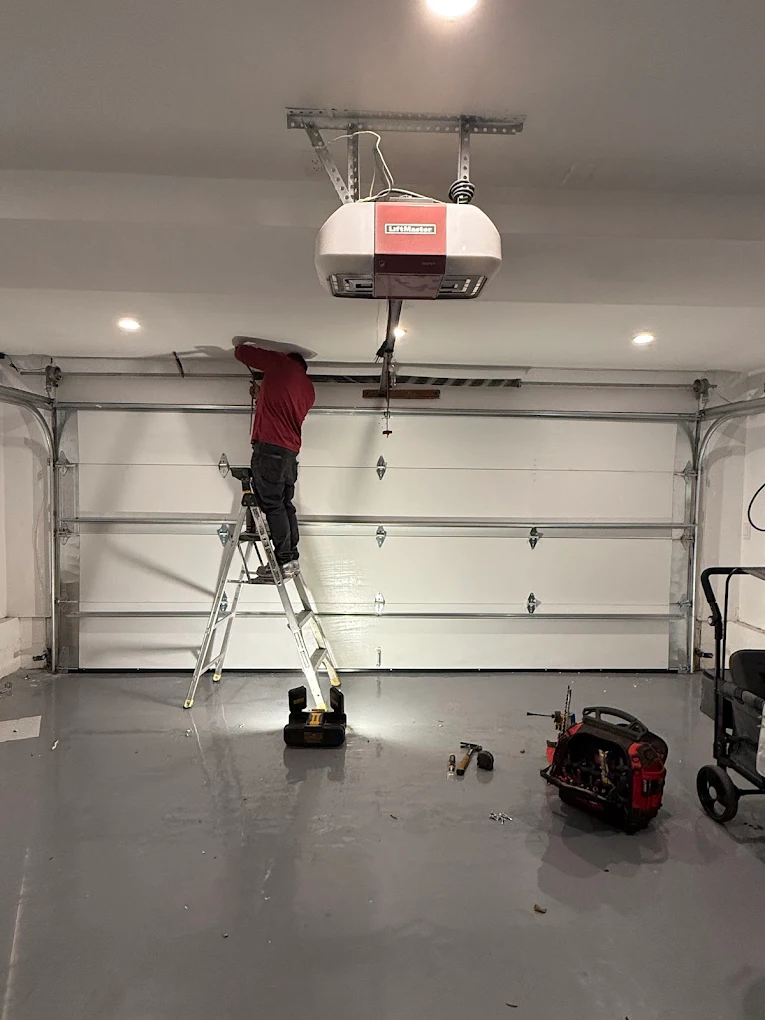 A technician standing on a stepladder to inspect the upper tracks and hardware of a white sectional garage door, emphasizing the importance of annual garage door inspections.