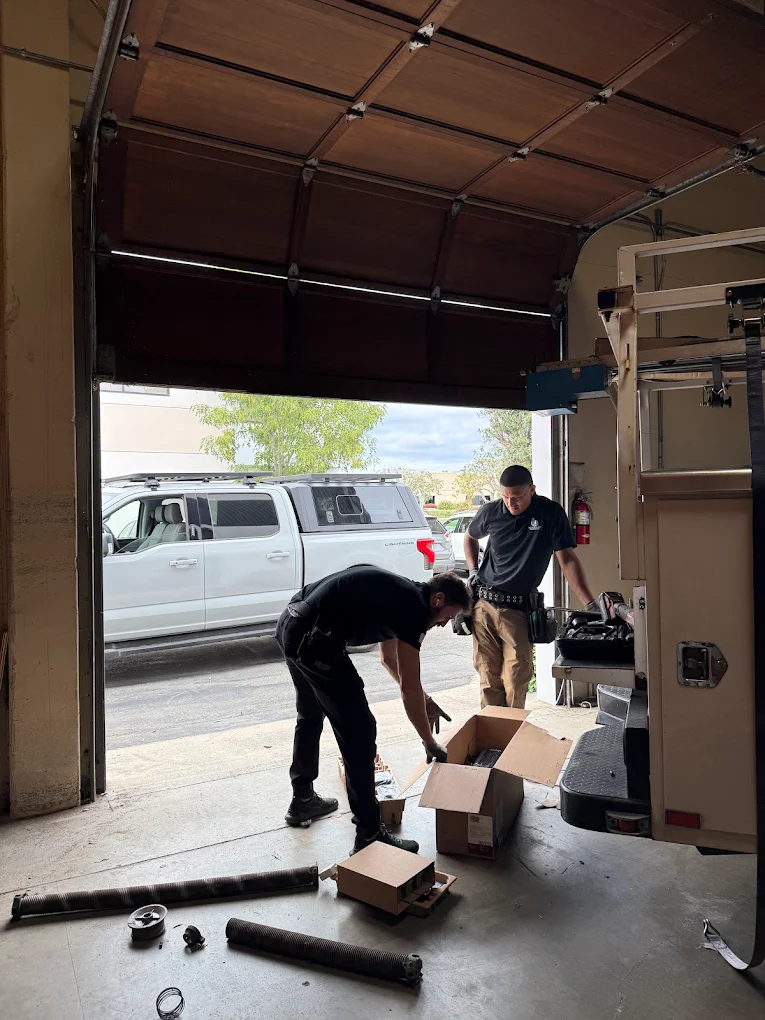 Two professional technicians preparing tools and replacement parts inside a garage with an open overhead wooden door, demonstrating essential safety precautions for garage door tune-ups.