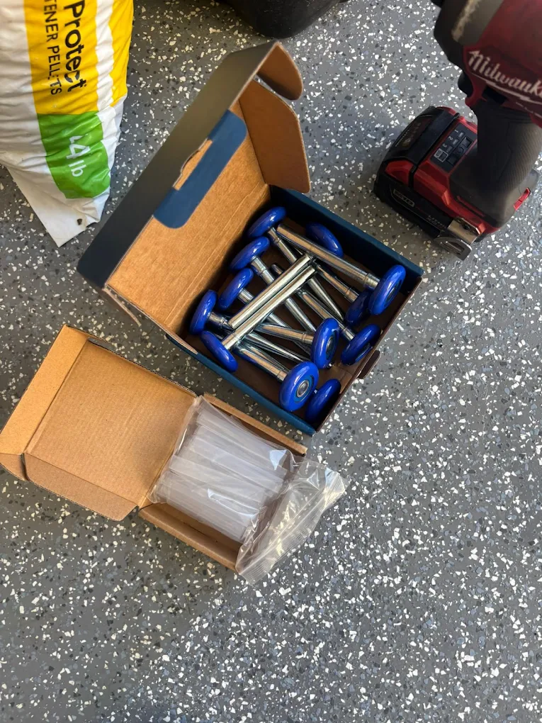 A cardboard box containing new blue nylon garage door rollers next to a package of plastic sleeves and a power drill, showing the replacement parts ready to install when garage door rollers are causing the noise.