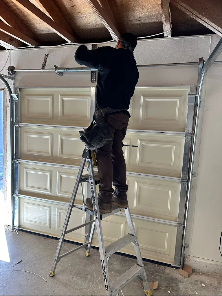 A professional technician standing on a stepladder to inspect and adjust the high-tension torsion spring assembly above a beige sectional garage door, highlighting the careful execution of a professional garage door inspection in Irvine, CA.