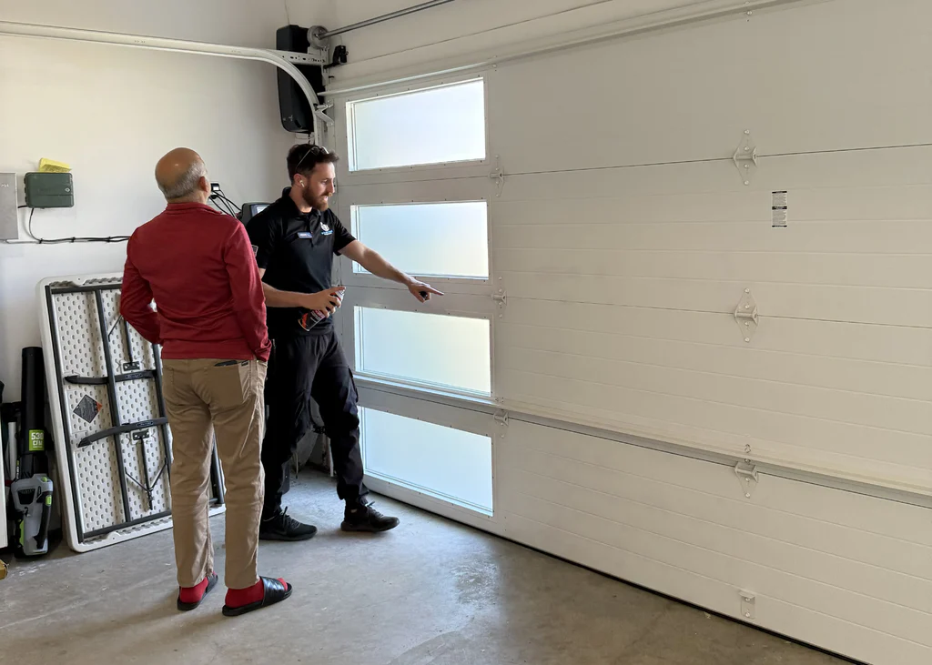A professional technician holding a spray can points out specific moving hardware pieces to a homeowner inside a garage, explaining what to check when a garage door begins to squeak.