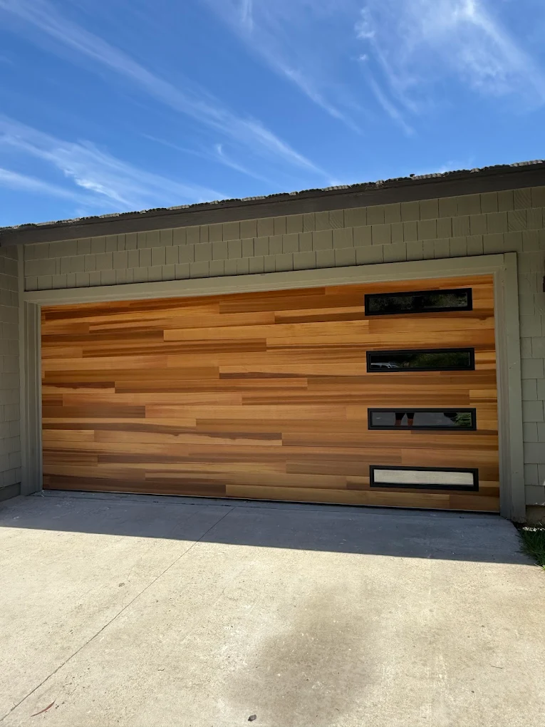 A modern sectional garage door featuring horizontal natural wood planks and a vertical row of small tinted rectangular windows on the right, showcasing a contemporary garage door style.